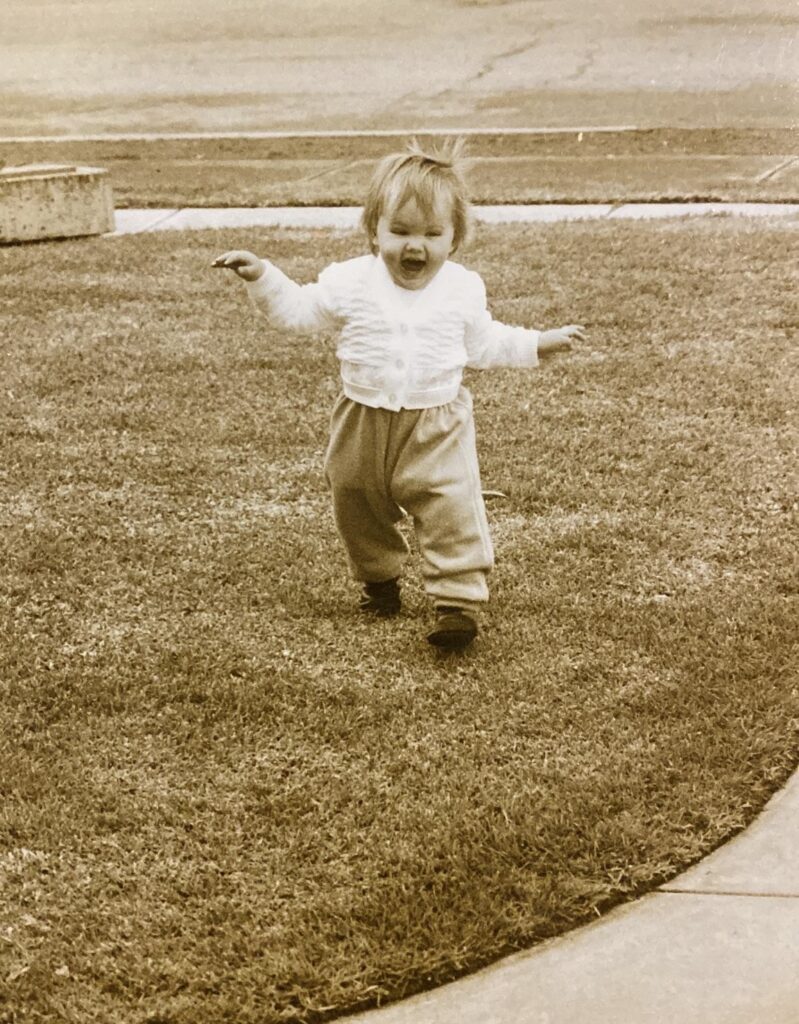 A toddler (me) runs across some grass, with a huge laughing smile and odd T-Rex arms.